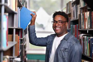 Dark-skinned handsome guy smiling camera, taking book in blue cover from the shelf with his hand. Close up smiling face young smart professor African nationality in university library, choosing book.