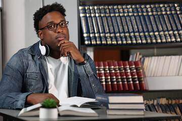 Pensive African male entrepreneur ponders in the office of a modern office at his desk. Young handsome African-American teacher sits in a classroom among books, holding his chin with his hand