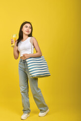Beautiful confident Asian woman stylishly dressed with a striped bag on her shoulder looks away and smiles. On a yellow background, a charming girl in a white T-shirt and jeans