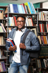 Confident african man in glasses and with books in hand on the background of shelves with books. Handsome young african curly-haired guy student in denim jacket with backpack in university library. 