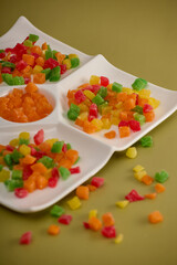 Sweet dessert in form candied fruit cubes close-up in a white menager on a yellow background. On yellow background, close-up dessert of colored candied fruit is laid out on white square-shaped plate. 