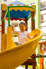 Una linda niña de dos años sonriente juega en un tobogan dentro del patio de un condominio. - obrazy, fototapety, plakaty