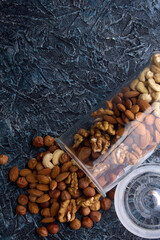 Mixture of different nuts is scattered on a gray table from a glass jar with an open lid. Nut mixture is scattered from a transparent glass jar on a gray textured background.