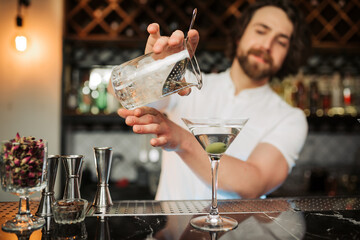 A skilled male bartender confidently pours a martini into a glass at a well-equipped, modern bar,...