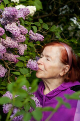 Outdoor close-up portrait of old woman. Beautiful elderly woman against background of blooming lilacs in spring park. Elderly lady woman enjoying scent of spring flowers in her garden