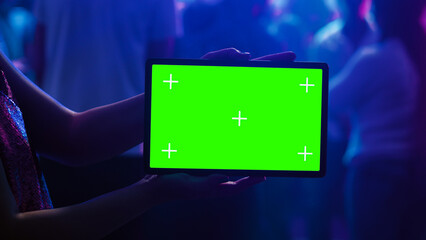 Adult using tablet with green screen at club, holding gadget with blank mockup template on dance floor. Young woman showing isolated display with chroma key and copyspace at nightclub.