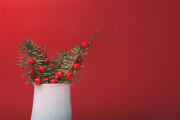 Bouquet of nobilis fir and winterberry branches in a vintage vase in front of red background....