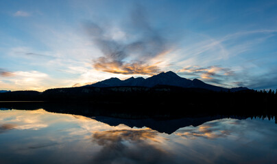 Sunset colored clouds and mountain are reflecting in a calm lake creating a peaceful scene.
