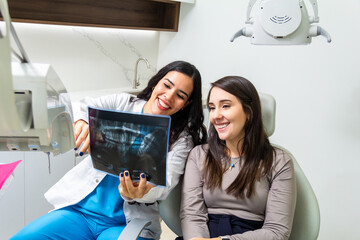 Obraz na płótnie Canvas Latin female smiling dentist explaining teeth x-ray scan to caucasian woman patient sitting in a dental chair. Health care concept.