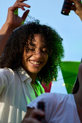 Vertical photo. Happy young African adult afro woman dancing with friends at rooftop party. Excited people have fun drinking beer outdoor in dusk. Lively Millennial female celebrating summer vacation.