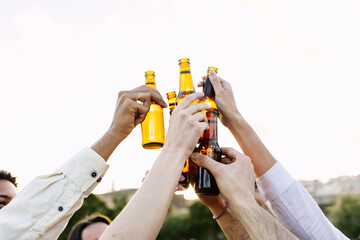 Group of young people celebrating together clinking beer bottles at summer party outdoors. Youth and beverage concept.