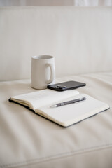 Vertical image of notebook, pen, phone and mug on a shite couch. Work from home