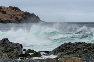 A stormy sea on a bright sunny day.  The ocean is a deep blue, coral green with large white waves. The coastline has ice and snow collecting among its rocks. The sky is blue with a thick ocean spray.