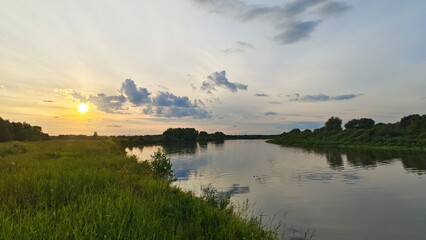 On a summer evening, the sun sets over the horizon and paints the sky and clouds in bright colors. Ripples on the water Tall grass, bushes and trees grow along the banks and are reflected in the water