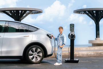 Little boy recharging eco-friendly electric car from EV charging station. EV car road trip travel by the seashore by alternative vehicle powered by clean renewable and sustainable energy. Perpetual - obrazy, fototapety, plakaty