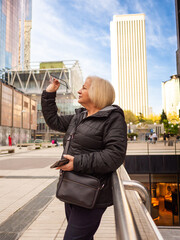 senior blonde business woman looks at her glasses with cell phone in hand in winter in office district - 789722914