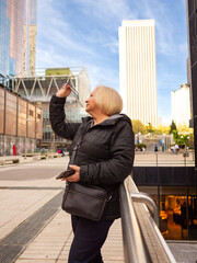 senior blonde business woman looks at her glasses with cell phone in hand in winter in office district - 789722966