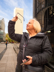 senior blonde business woman looks at her glasses with cell phone in hand in winter in office district - 789723114