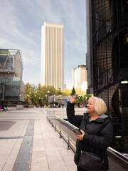 senior blonde business woman looks at her glasses with cell phone in hand in winter in office district - 789723180