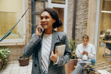 Smiling business woman standing at office terrace and talking by phone on colleague background