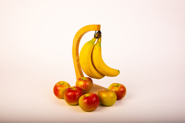Bananas hanging from stand with apples around base on white seamless background.