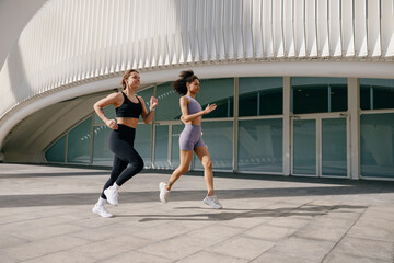 Two young women in sportswear are running on modern buildings background. Active lifestyle concept