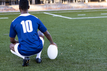 African American young male athlete sitting on grass, holding a rugby ball, copy space