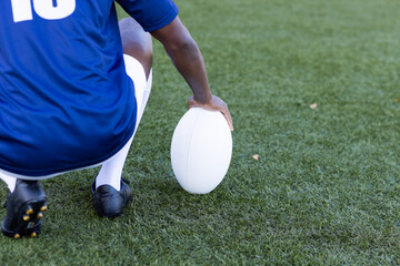 An African American young male athlete wearing a blue jersey is holding a rugby ball on a grass fiel