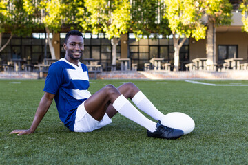 African American young male athlete sitting on grass, holding a rugby ball, copy space