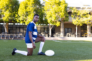 African American young male athlete kneeling on field outdoors, holding a rugby ball, copy space