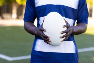African American young male athlete holding a rugby ball, wearing a blue and white jersey, on field 