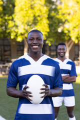 Two African American young male athletes are holding a rugby ball, smiling, on a field outdoors