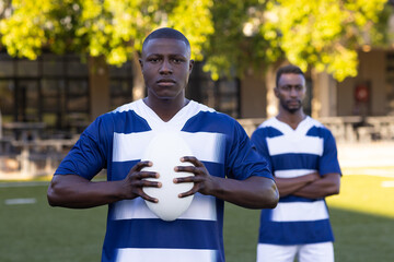 Two African American young male athletes are holding a rugby ball on a field outdoors