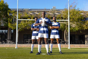 Three African American young male athletes holding a rugby ball on a field outdoors