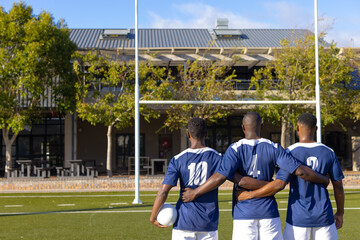 Three African American young male athletes holding a rugby ball, standing on a field, copy space