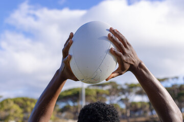 African American young male athlete, on field outdoors, hands holding rugby ball high against a sky 