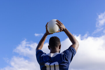 African American young male athlete holding white rugby ball above head on field, copy space