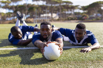 Three African American young male athletes reaching for a rugby ball on a grass field