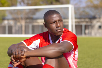 African American young male athlete sitting on grass on field outdoors, looking thoughtful
