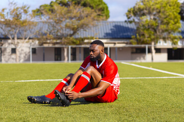 African American young male athlete sitting on grass outdoors, tying shoe laces, copy space