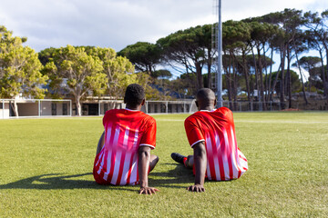 Two African American young male athletes sitting on grass, looking at a field outdoors