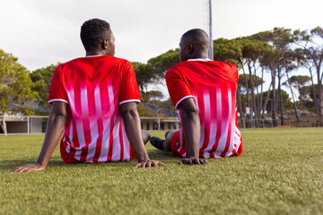 Two African American young male athletes are sitting on grass on field outdoors, wearing red jerseys
