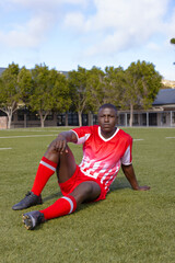 An African American young male athlete sitting on grass on field outdoors, wearing soccer gear