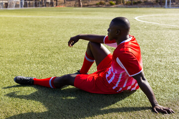 African American young male athlete sitting on grass on field outdoors, wearing red soccer uniform