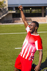 African American young male athlete in red & white jersey celebrates on field outdoors, copy space