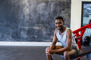 African American young male athlete sitting on a bench in a gym, smiling, copy space