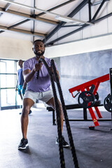 Obraz premium Two African American young male athletes are training with battle ropes in gym