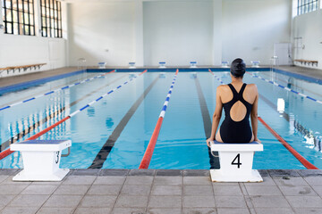 Biracial young female swimmer sitting at pool edge indoors, looking at water, copy space