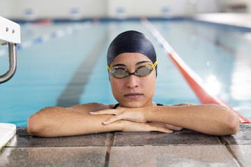 Young biracial female swimmer resting at pool edge indoors, wearing goggles