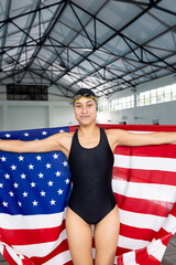 Biracial young female swimmer standing indoors wrapped in American flag, smiling at camera